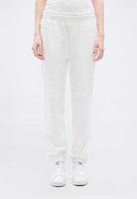 Άτομο που φοράει λευκά τζόγκερ με ελαστική μέση και λευκά παπούτσια Lacoste, στέκεται μπροστά σε ένα απλό φωτεινό φόντο.