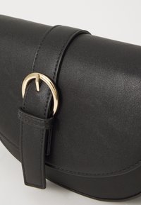 Bolso de mano de cuero sintético negro con un diseño curvado, que cuenta con una hebilla de anillo en D en tono dorado y una correa, textura suave y acentos minimalistas.