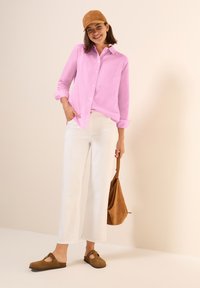 Camicia button-up rosa chiaro, pantaloni bianchi corti, scarpe in suede marrone con ritagli, borsa in suede marrone, cappellino beige. Look semplice e casual.