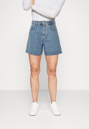Persoon draagt middenblauwe denim shorts, witte longsleeve en witte sneakers staand tegen een effen witte achtergrond.