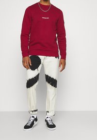 Sweat-shirt rouge avec logo, associé à un jean blanc avec des motifs tie-dye noirs. Baskets noires avec semelle et lacets blancs.
