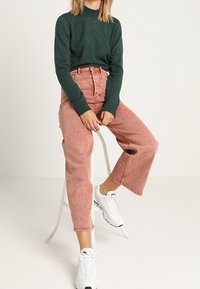 Dark green knit sweater paired with high-waisted, light pink denim jeans. White sneakers feature a textured sole. Individual seated on a stool.