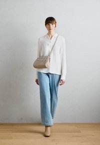 Beige crossover taske med rektangulær form, lavet af glat materiale, med en frontlomme og justerbar rem. Båret med casual lyseblå jeans og hvid skjorte.