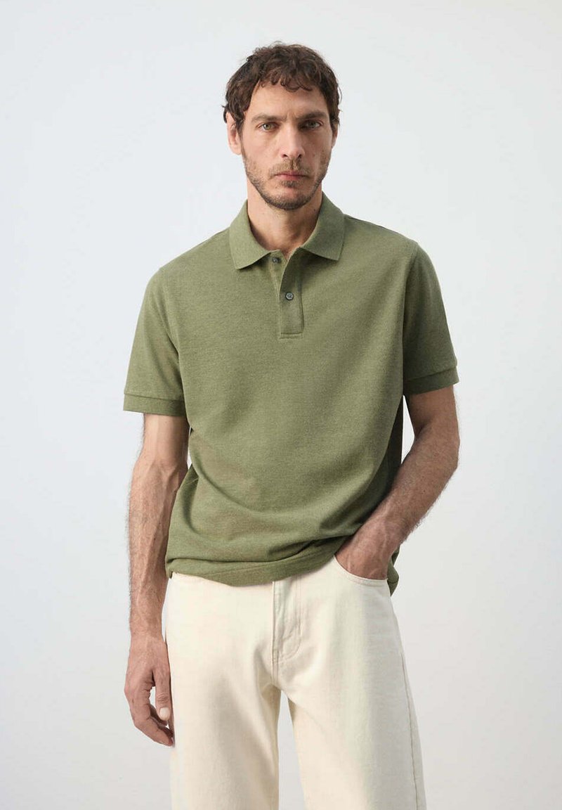 Homme portant un polo manches courtes vert olive et un pantalon crème, debout avec une main dans la poche devant un fond uni.