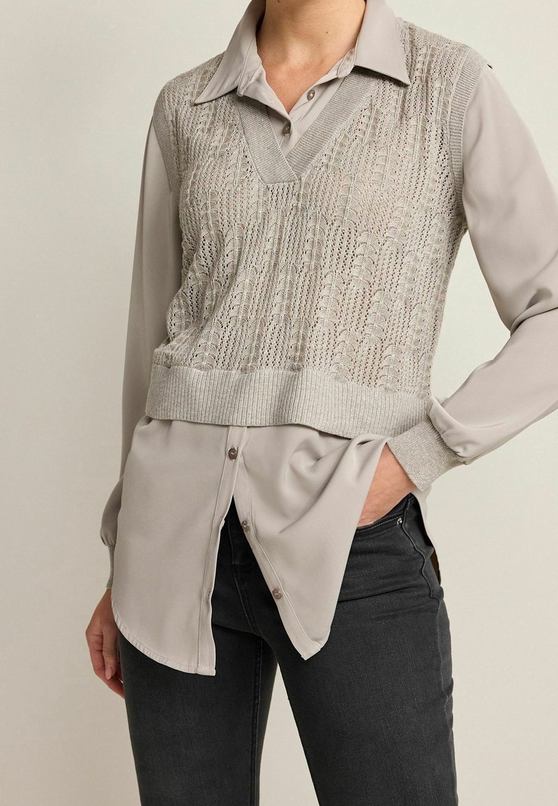 Gilet grigio chiaro lavorato a maglia sopra una camicia grigia con bottoni, con scollo a V e motivo testurizzato; indossato con jeans grigi scuri.