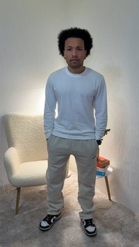 Pessoa vestindo uma camisa branca de manga comprida e calças de fato de treino cinza claro, em pé sobre um tapete, com ténis preto e branco, contra uma parede clara.