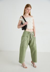 Ung kvinde iført hvid off-shoulder top, løse olivengrønne bukser med store lommer, hvide strophæle og en sort skuldertaske.