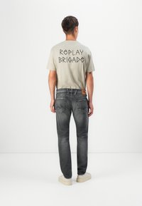 Light grey t-shirt with "REPLAY BRIGADE" in black text on the back, paired with dark grey jeans and cream sneakers. Simple design, casual fit.