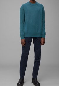 Tyrkysový pletený svetr s kulatým výstřihem a dlouhými rukávy, v kombinaci s tmavě námořnickými manšestrovými kalhotami a černými kotníkovými botami, na neutrálním šedém pozadí.