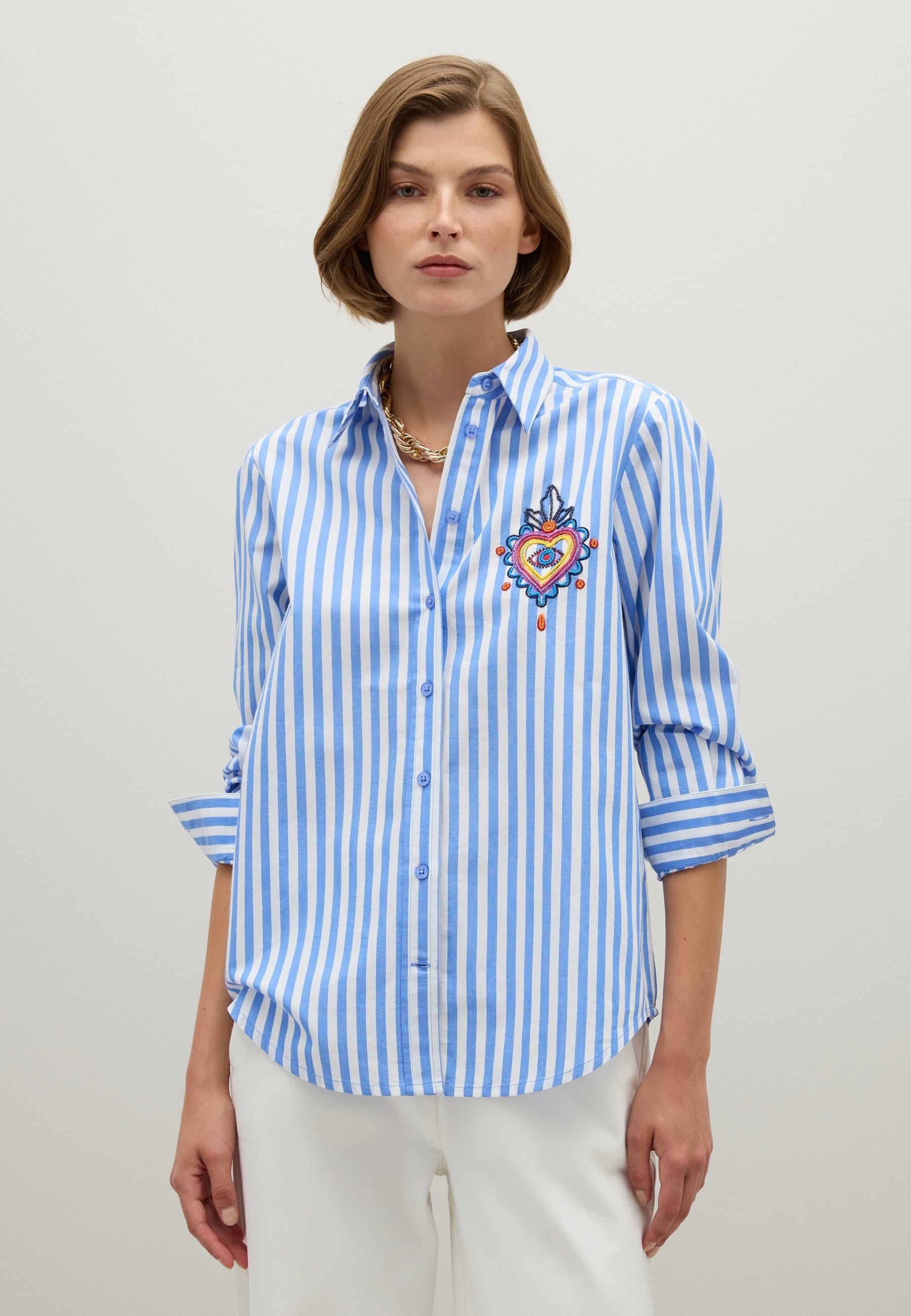 Woman wearing blue and white vertical striped button-up shirt with colorful embroidered heart and white pants, standing against plain background.