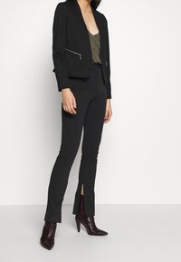 Blazer noir sur mesure avec poches zippées, associé à un pantalon noir ajusté avec des fentes latérales. Porté avec des bottines bordeaux à bout pointu.