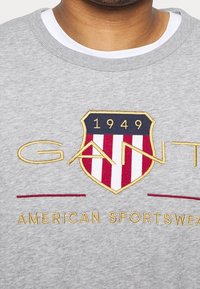 Felpa grigia con ricamo a scudo in rosso, bianco e blu. Riporta la scritta "GANT" e "AMERICAN SPORTSWEAR" in oro.