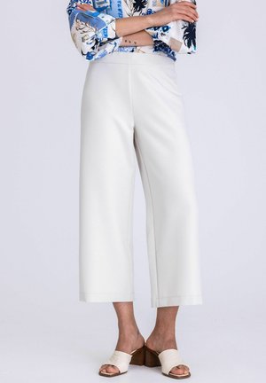 Vrouw met witte, wijde cropped broek en witte open-toe blokhak sandalen, armen gekruist, tegen een effen lichte achtergrond.