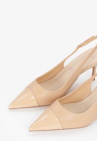 Talons slingback beiges avec bouts pointus, présentant un capuchon en verni brillant et une tige en cuir lisse. Sangle de cheville réglable incluse.