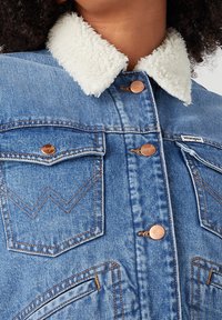 Jeansjacka i ljusblå med en vit fuskpäls-krage. Har kopparknappar och dubbla bröstfickor med chevron-stygn.