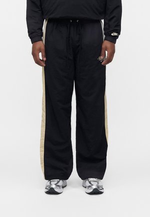 Person iført sorte Nike træningsbukser med beige sidelister, en sort sweatshirt med Nike-logo på ærmet og hvide løbesko.