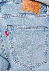 Jean en denim bleu clair avec une poche arrière, une étiquette en tissu rouge et un patch en cuir marron indiquant la marque et les détails de taille.