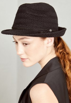 Kobieta o jasnej karnacji i czerwonych włosach związanych do tyłu, nosząca czarny pleciony kapelusz fedora i czarny top bez rękawów, patrząca w dół i na bok.