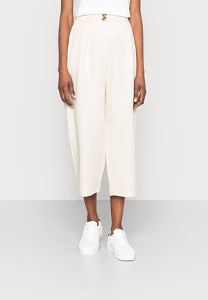 Kobieta ubrana w kremowe spodnie z wysokim stanem i szerokimi nogawkami, zapinane na dwa guziki, w połączeniu z białymi trampkami i włożonym do środka białym topem.