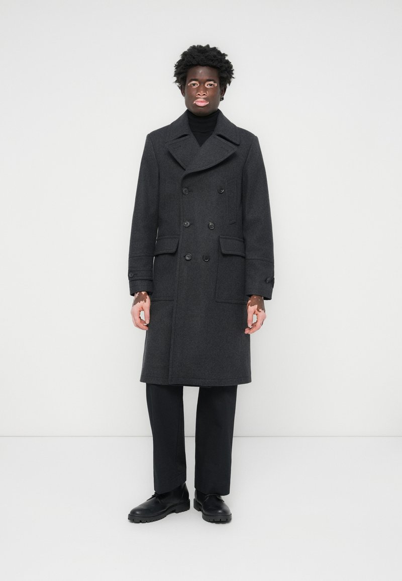Manteau double boutonnage gris foncé, en mélange de laine, avec un large col à revers, deux poches avant, fermetures à boutons, associé à un pantalon noir et des chaussures.