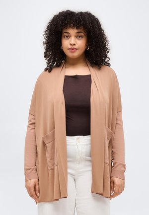 Femeie cu păr creț, purtând cercei aurii în formă de cerc, un cardigan bej drapat, un top închis la culoare și pantaloni albi, stând în picioare pe un fundal simplu.