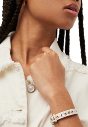 Gros plan sur une personne aux cheveux tressés portant une veste blanche et un bracelet blanc à œillets métalliques, tenant son col.