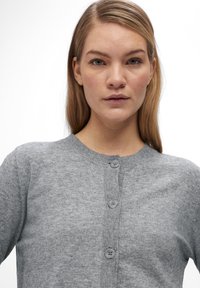 Grijze gebreide cardigan met een ronde halslijn, voorzien van een knopen sluiting en geribbelde details langs de knopenlijst en mouwen. Zachte textuur.