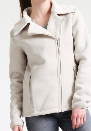 Nainen pukeutuneena vaaleanbeigeen fleece-takkiin, jossa on pehmeä sherpa-vuorattu kaulus, vetoketjullinen etuosa ja vetoketjulliset sivutaskut, yhdistettynä tummiin farkkuihin.