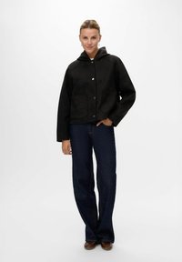 Giacca nera con cappuccio e bottoni a pressione, due tasche frontali e vestibilità rilassata, abbinata a jeans svasati blu scuro. Design minimalista.