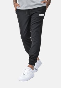Černé joggingové kalhoty s elastickým pasem na šňůrku, bočními kapsami a bílou nášivkou s logem. Spárované s bílými teniskami s perforovanými detaily.