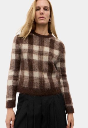 Kvinde iført en brun og cremefarvet ternet fuzzy sweater kombineret med sorte plissébukser mod en ensartet lys baggrund.