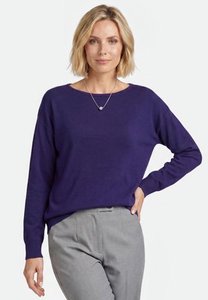 Kvinde med kort blondt hår iført en lilla langærmet sweater, grå bukser og en sølvhalskæde med rund vedhæng.
