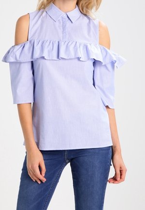 Vrouw die een lichtblauwe cold-shoulder blouse draagt met ruches over de borst en een donkerblauwe spijkerbroek, tegen een effen achtergrond.