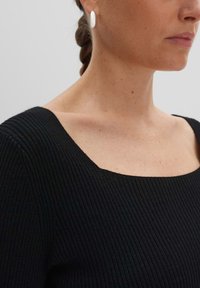 Schwarzes, geripptes Top mit quadratischem Ausschnitt. Glatte Textur mit figurbetontem Design. Weiße ovale Ohrringe im Ohr setzen einen Kontrast.