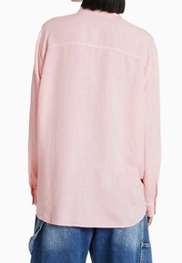 Light pink long-sleeve shirt with a relaxed fit, featuring a back seam detail and buttoned cuffs, paired with loose blue jeans.