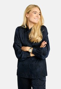 Tumši zila, garām piedurknēm blūze ar teksturētu č�ūskas rakstu. Ar apkakli un pogām piedurkņu galiem, kopā ar atbilstošām biksēm.