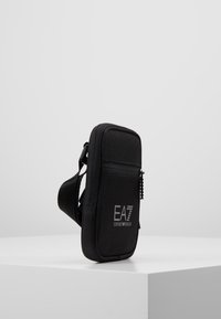 Borsa a spalla in tessuto nero con chiusura a zip, forma rettangolare, caratterizzata da un logo sulla parte frontale e una tracolla regolabile per il trasporto.