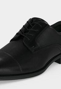 Chaussure formelle en cuir noir avec lacets, bout arrondi et détails cousus à l'avant et sur le côté, présentée sur un fond clair uni.