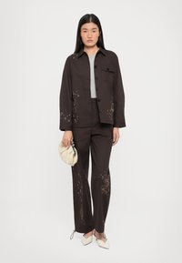 Brown embroidered jacket and pants set with floral and fish designs, paired with a light grey top and cream clutch bag. Square-toed shoes.