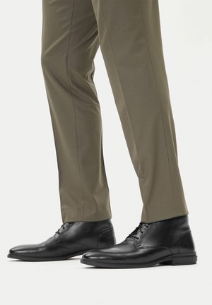 Črne usnjene elegante čevlje z gladko teksturo in vezalkami, združene z lahkimi olivnimi ozkimi hlačami. Klasična silhueta in subtilen lesk.