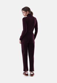 Burgunderfarbenes Jumpsuit mit Stehkragen, tailliertem Oberteil und angepassten Hosen; mit Schlitzen am Saum und kombiniert mit weißen und burgunderfarbenen Schuhen.
