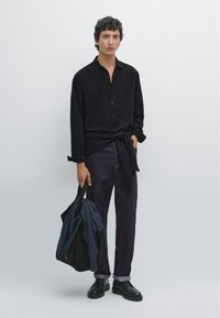 Schwarz geknöpftes Hemd, marineblaue Hose und schwarze Schuhe. Trägt eine dunkle Tragetasche mit gefalteter Kleidung. Glatte Textur, lockere Passform, minimalistisches Design.