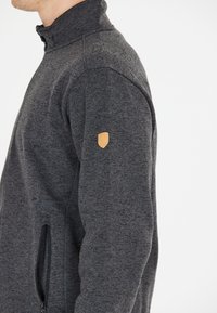 Grå fleecejakke med høj krave, struktureret overflade, lynlås side lomme og et lille logo-patch i tan læder på ærmet.
