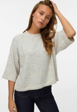 Young woman with long wavy hair wearing beige loose three-quarter sleeve sweater and dark jeans, posing with one hand touching her head.