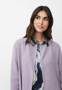 Ljuslila cardigan med en texturerad stickad design, bärs över en blå och vit marmorerad skjorta med krage och knapppanel.