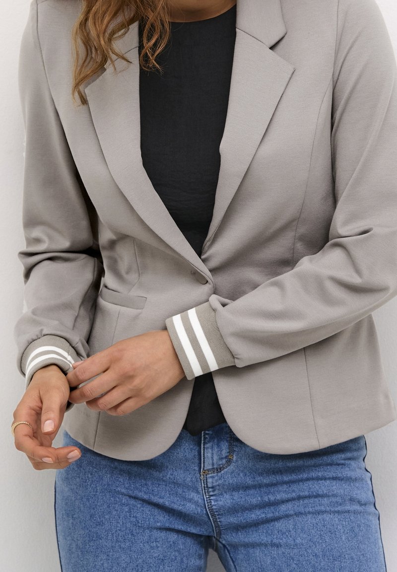 Kvinde justerer manchetterne på en beige blazer med hvide stribede ribbede manchetter, iført sort top og blå jeans.