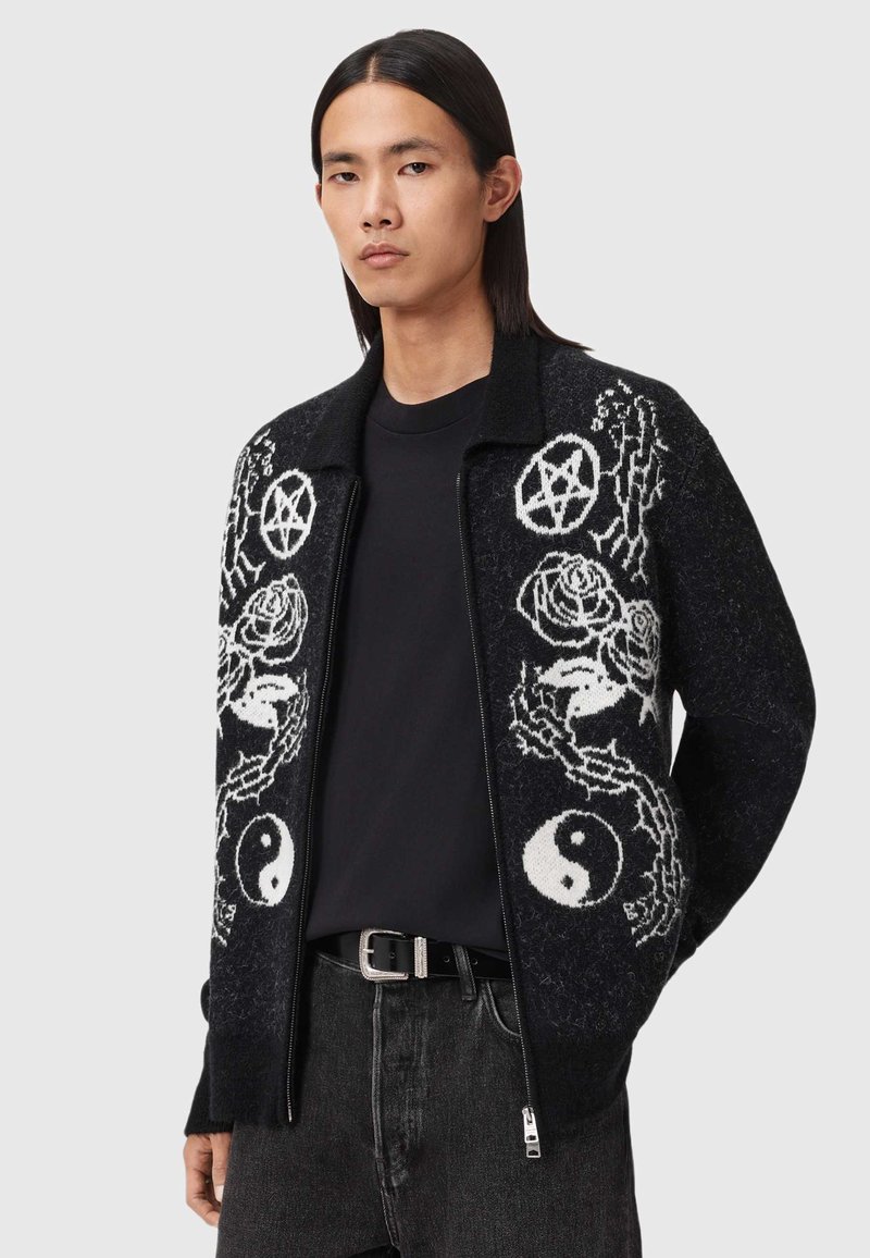 Zwarte gebreide cardigan met ritssluiting aan de voorkant, versierd met witte patronen van rozen, yin-yang symbolen en pentagrammen. Geribbelde manchetten en kraag.