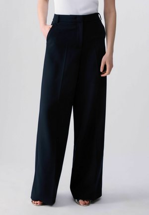 Nő, aki fekete, magasderekú, széles szárú nadrágot visel elülső redőkkel, ezüst nyitott orrú magassarkúval és fehér ujjatlan felsővel párosítva.