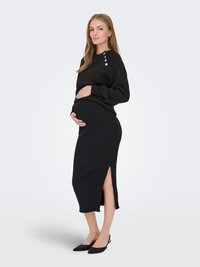 Nėščia moteris juodu megztiniu su sagomis ant peties ir juodu ribuotu sijonu su šoniniu skeltuku, stovinti ir laikanti savo pilvą.