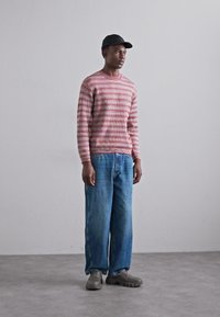 Pulover tricotat cu dungi roșii și albe, blugi lejeri albaștri și saboți gri, purtați cu o șapcă neagră. Design simplu, cu o croială relaxată și textură.
